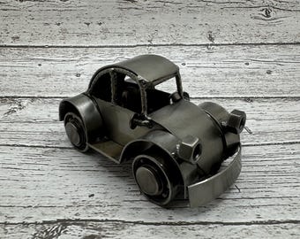 May include: A silver metal model car shaped like a classic Volkswagen Beetle. The car is sitting on a white wooden surface.
