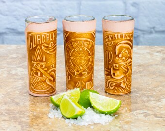 May include: Three brown leather-wrapped shot glasses with different designs. The glasses are on a light brown surface with lime wedges and salt.
