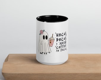 May include: White ceramic mug with a black interior and a cartoon ghost illustration. The ghost has a pink flower and is holding a coffee cup. Text on the mug reads "Hocus Pocus I Need Coffee To Focus."