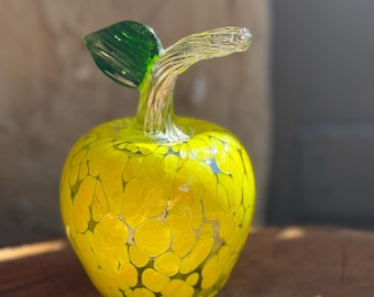May include: A hand-blown glass apple sculpture featuring a bright yellow body with a speckled, translucent pattern. The apple is topped with a clear, textured glass stem and a single, smooth, vibrant green glass leaf attached at the top.