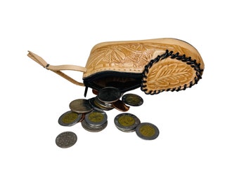 May include: A tan leather coin purse with intricate floral designs is open, spilling out silver coins. The purse has a zipper closure and a leather strap.