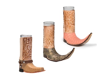 May include: Three leather shot glasses shaped like cowboy boots. The boots are decorated with brown and tan leather carvings. The boots are on a white background.