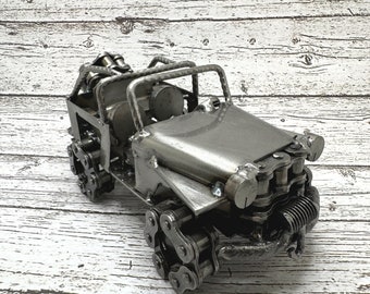 May include: A handcrafted metal sculpture of a jeep. The sculpture is made from various metal pieces, including chains for the wheels and other components. The jeep has a roll bar and a detailed engine. The metal has a silver finish.