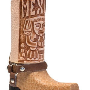 Puede incluir: Un vaso de chupito de cuero marrón con forma de bota vaquera con la palabra "MEXICO" en relieve en el lateral. La bota tiene una correa de cuero marrón con una hebilla de metal y un inserto de vidrio.