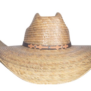 K&ouml;nnte beinhalten: Ein breitkrempiger Strohhut im Cowboy-Stil mit einem braunen Lederband und silbernen Akzenten. Der Hut hat eine nat&uuml;rliche, hellbraune Farbe.