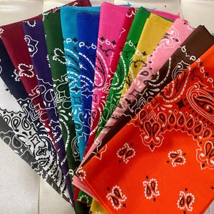May include: A collection of colorful bandanas, each folded to display a corner with a paisley pattern. The bandanas are in various colors, including red, blue, green, pink, and yellow, with white or black paisley designs. The bandanas are made of a soft fabric.