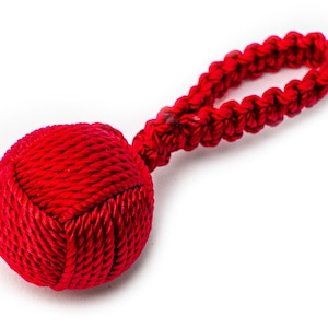 Può includere: Una palla rossa di corda con un manico ad anello. La palla è fatta di corda rossa tessuta strettamente e il manico è fatto della stessa corda, ma intrecciata.