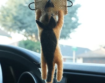 May include: A brown and black felt dog hanging from a rearview mirror in a car. The dog is hanging upside down and has a burlap tag around its neck.