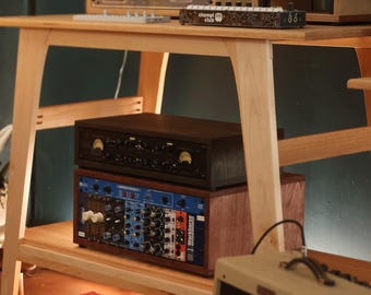 May include: A light-colored wooden studio desk with a lower shelf, holding various audio equipment including a black synthesizer, a wooden-cased unit, a black amplifier with analog meters, and a blue modular synthesizer rack with multiple knobs.
