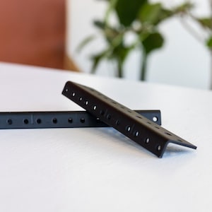 May include: Two black metal shelf brackets with holes for mounting. The brackets are angled and have a flat surface.