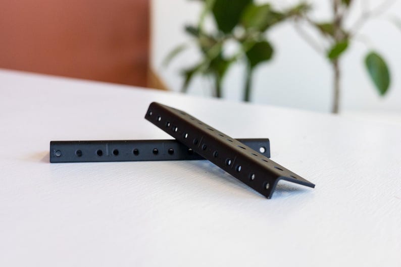 May include: Two black metal shelf brackets with holes for mounting. The brackets are angled and have a flat surface.