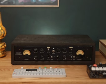 May include: A dual-channel audio compressor housed in a dark wooden enclosure with a black front panel. It features two analog VU meters, multiple rotary control knobs labeled for attack, release, input, and output, and a central power toggle switch.