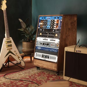 Op de afbeelding: Een houten rack-studioconsole op wielen met diverse audioprocessors, waaronder een Cloudlifter CL-4 Mic Activator en een QSC-versterker, geplaatst naast een lichtgekleurde V-vormige elektrische gitaar en een beige versterker.