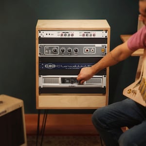 Op de afbeelding: Een houten rackbehuizing met meerdere audiocomponenten, waaronder een zilveren QSC-eindversterker, een blauwe Cloudlifter CL-4 microfoonactivator, een grijze voorversterker en een rack-interface met diverse knoppen en ingangspoorten.
