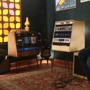 Op de afbeelding: Twee houten studiostandaards met zwarte metalen haarspeldpoten. De linkerstandaard bevat diverse audioapparatuur met blauwe en zwarte panelen. De rechterstandaard is voorzien van een Cloudlifter-unit en een QSC-versterker in een licht houten frame.