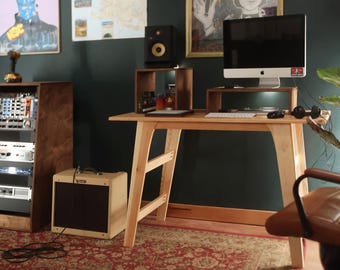 May include: A light-colored wooden desk with angled legs and a horizontal support brace. The desk surface holds a computer monitor on a wooden riser, a keyboard, and a mouse. A beige guitar amplifier sits on the floor to the left of the desk.