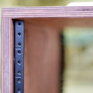 May include: Close-up of a wooden cabinet with a black metal rack mounted inside. The cabinet is made of plywood and has a natural wood finish.