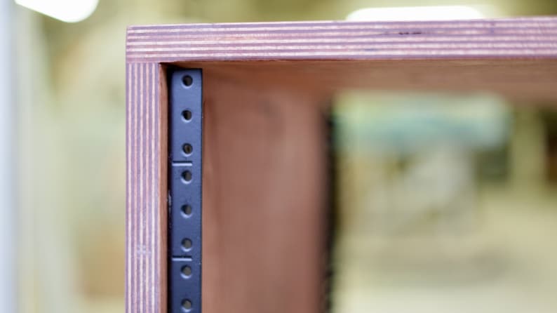 May include: Close-up of a wooden cabinet with a black metal rack mounted inside. The cabinet is made of plywood and has a natural wood finish.