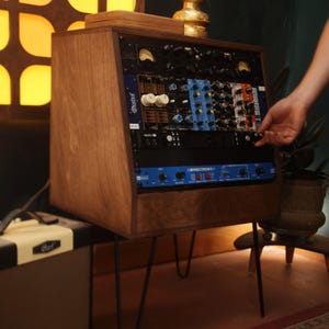 Op de afbeelding: Een houten studiokast met een schuine voorzijde, ondersteund door vier dunne zwarte metalen haarspeldpoten. Het rack bevat meerdere audioverwerkingseenheden, waaronder een blauwe Effectron II-unit, een Apollo-interface en diverse modules.