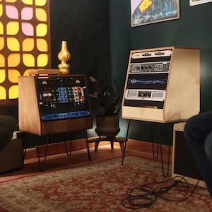Op de afbeelding: Twee houten studiostandaards met zwarte metalen haarspeldpoten. De linkerstandaard bevat diverse audioapparatuur met blauwe en zwarte panelen. De rechterstandaard is voorzien van een Cloudlifter-unit en een QSC-versterker in een licht houten frame.