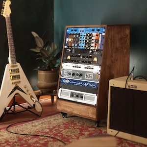 Op de afbeelding: Een houten rack-studioconsole op wielen met diverse audioprocessors, waaronder een Cloudlifter CL-4 Mic Activator en een QSC-versterker, geplaatst naast een lichtgekleurde V-vormige elektrische gitaar en een beige versterker.