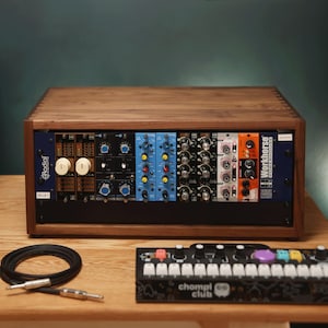 Op de afbeelding: Een houten rack met meerdere audiomodules, waaronder Radial- en Workhorse-units met diverse knoppen en schakelaars. Daarnaast liggen een zwarte audiokabel en een Chompi Club synthesizer met een toetsenbord en kleurrijke knoppen.
