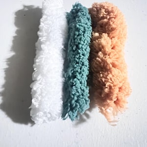 May include: Three fluffy, textured, cylindrical objects in white, teal, and peach colors are arranged vertically against a white background. The objects appear to be made of a soft, looped material, possibly yarn or fabric.