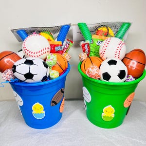 May include: Two colorful Easter baskets filled with sports-themed toys and treats. One blue bucket features baseball, football, and basketball decorations, while the other is green. Each basket contains a baseball, soccer ball, and orange basketball-shaped eggs.