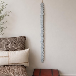 May include: A vertical wall hanging composed of a long string of circular, light gray felt discs. The discs are connected in a single column, suspended from a thin, light-colored cord attached to the top, creating a linear, textured decorative element.