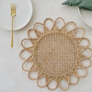 May include: A round, woven placemat with a sunburst design. The placemat is made of natural-colored rattan and features a central woven pattern. A gold fork and white plate are visible on the left. A green napkin and white plate are on the right.