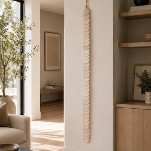 May include: A vertical wall hanging consisting of a long string of light-colored, circular wooden discs. The discs are arranged in a single column, overlapping slightly, and are suspended from a thin cord attached to the top of the wall.