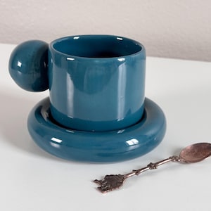 May include: A teal blue ceramic teacup with a unique round handle and matching saucer. A small silver spoon is also included.