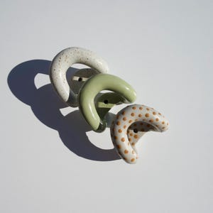 Può includere: Tre ganci da parete in ceramica a forma di U disposti in diagonale. Il primo è bianco sporco con macchie marroni, il secondo è verde salvia a tinta unita e il terzo è bianco sporco con un motivo a piccoli pois marroni uniformemente distanziati.