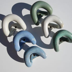 Pode incluir: Um conjunto de seis ganchos de parede em cerâmica com um formato curvo de ferradura. Os ganchos apresentam um acabamento liso e brilhante em várias cores, incluindo branco sujo, verde sálvia e azul claro, cada um com um orifício central.