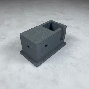 May include: A gray 3D printed model of a small building with a square base and a rectangular opening on one side. The building has two small square windows on the front.