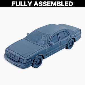以下が含まれることがあります： 完全に組み立てられた、青灰色のモデルカー。 ミニチュア車両は、窓、タイヤ、フロントグリルなど、詳細な成形が施されたセダンです。 テキスト「FULLY ASSEMBLED」は、画像の上部に白で印刷されています。