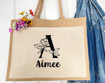May include: A natural jute tote bag with a white canvas front. The bag has a black floral design with the letter "A" and the name "Aimee" printed on the front.