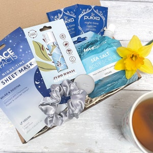 May include: A gift box filled with self-care items, including a blue sheet mask, a blue and green lip balm, a grey hair scrunchie, a white tea light candle, a blue sea salt body scrub, and two blue tea bags. The box is sitting on a white wooden surface next to a cup of tea and a yellow daffodil.