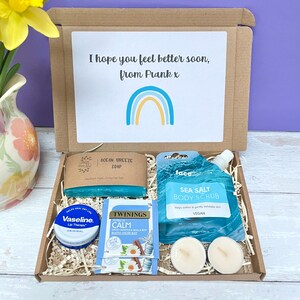 May include: A brown cardboard box filled with a self-care gift set. The box contains a blue soap bar, a blue body scrub, a lip balm, a tea bag, and two white tea lights. The box has a handwritten note that says "I hope you feel better soon, from Frank x" and a rainbow illustration.