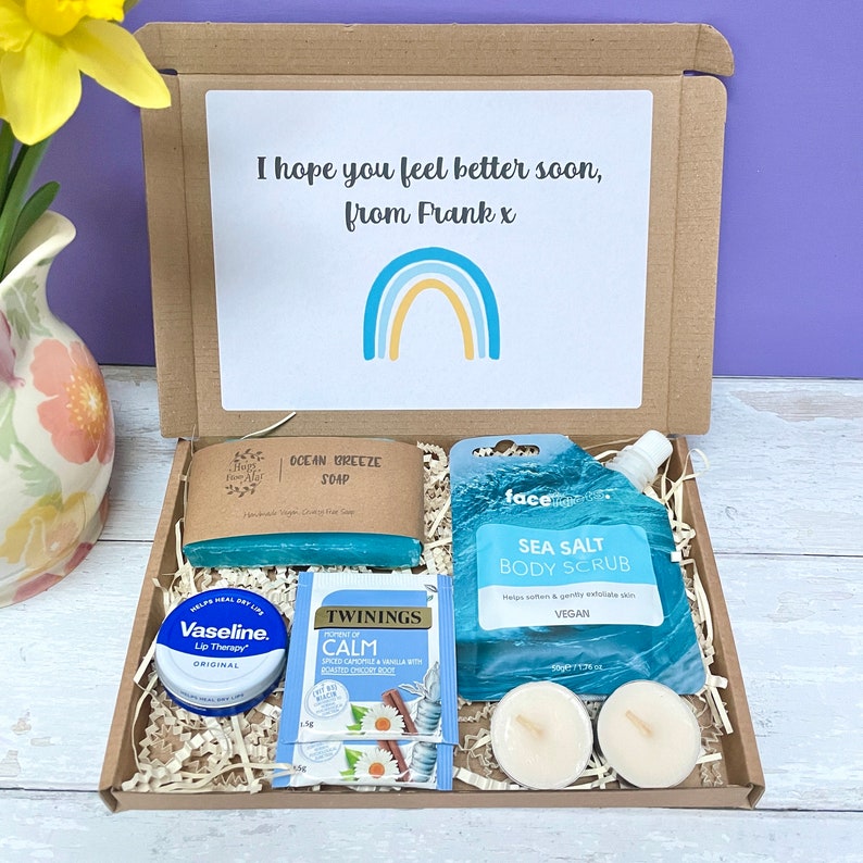 May include: A brown cardboard box filled with a self-care gift set. The box contains a blue soap bar, a blue body scrub, a lip balm, a tea bag, and two white tea lights. The box has a handwritten note that says "I hope you feel better soon, from Frank x" and a rainbow illustration.