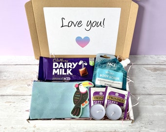 May include: A gift box filled with various items, including a chocolate bar, body scrub, tea bags, candles, and a small blue pouch with a toucan design. The box has a handwritten note that says "Love you!"