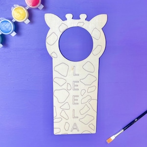 May include: Wooden giraffe-shaped door hanger with a cutout for a handle. The hanger is decorated with a giraffe print and the name "LEELA" is written vertically down the centre.