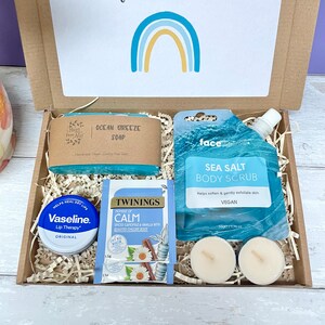 May include: A gift box filled with self-care items, including a blue soap bar labeled "Ocean Breeze Soap", a blue body scrub pouch labeled "Sea Salt Body Scrub", a small container of lip balm labeled "Vaseline Lip Therapy", a tea bag labeled "Twinings Moment of Calm", and two white tea lights.