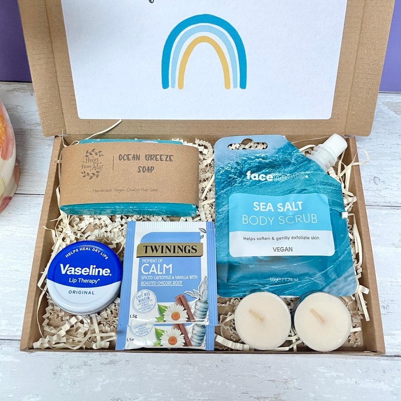 May include: A gift box filled with self-care items, including a blue soap bar labeled "Ocean Breeze Soap", a blue body scrub pouch labeled "Sea Salt Body Scrub", a small container of lip balm labeled "Vaseline Lip Therapy", a tea bag labeled "Twinings Moment of Calm", and two white tea lights.