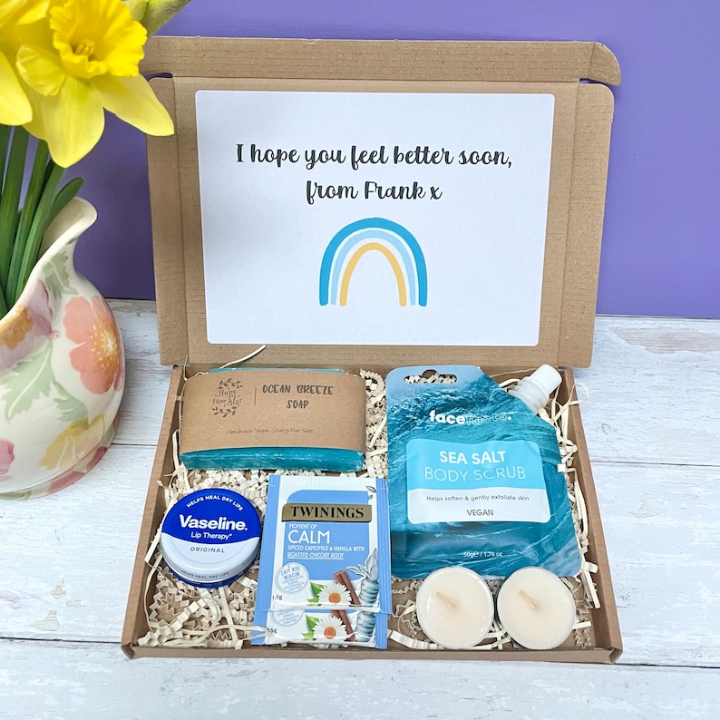 May include: A brown cardboard box filled with a get well soon gift set. The box contains a blue soap bar, a blue body scrub, a small container of lip balm, a tea bag, and two white tea lights. The box has a card on top that says "I hope you feel better soon, from Frank x" with a rainbow illustration.