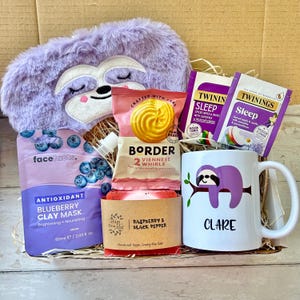 May include: A gift box containing a purple sloth eye mask, a mug with a sloth design and the name "Clare", blueberry clay mask, raspberry and black pepper soap, biscuits, and sleep tea bags. The box includes various self-care products.