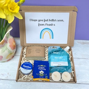 May include: A brown cardboard box filled with self-care items, including a bar of soap, a tube of body scrub, a small jar of lip balm, tea bags, and two tealight candles. The box has a card on top with the message "I hope you feel better soon, from Frank x" and a rainbow graphic.