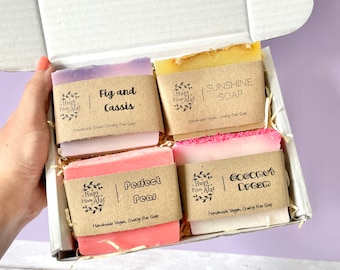 May include: A gift box filled with four bars of handmade vegan, cruelty-free soap. The soaps are labeled "Fig and Cassis", "Sunshine Soap", "Perfect Pear", and "Coconut Dream".