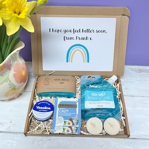 May include: A brown cardboard box with a blue and yellow rainbow design on the lid. The box contains a variety of self-care items, including a bar of soap, a body scrub, lip balm, tea, and candles. The box also includes a handwritten note that says "I hope you feel better soon, from Frank x".
