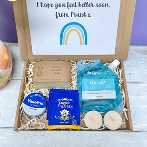 May include: A gift box filled with self-care items, including a bar of honey oatmeal soap, a tube of sea salt body scrub, a small tin of lip balm, two tea bags, and two tea lights. The box is decorated with shredded paper and a card that reads "I hope you feel better soon, from Frank x".